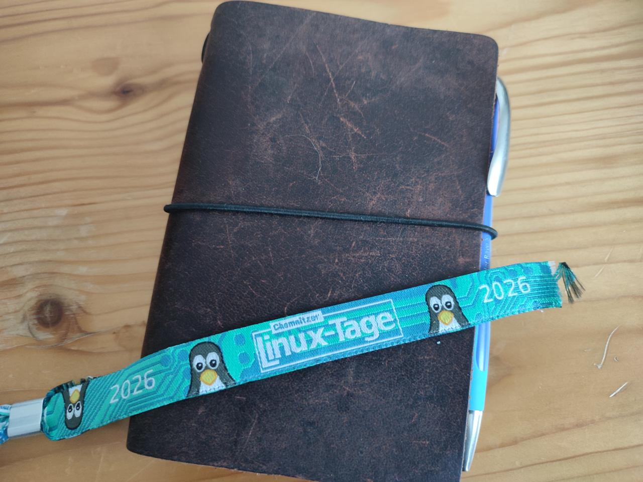 Auf einem in Leder eingebundenen Notizbuch liegt ein Armband mit der Aufschrift „Chemnitzer Linuxtage 2026“.