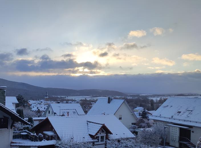 Ein Blick über die Dächer eines Dorfs im Winter. Die Dächer sind mit Schnee bedeckt. Die Sonne scheint durch die Wolken.
